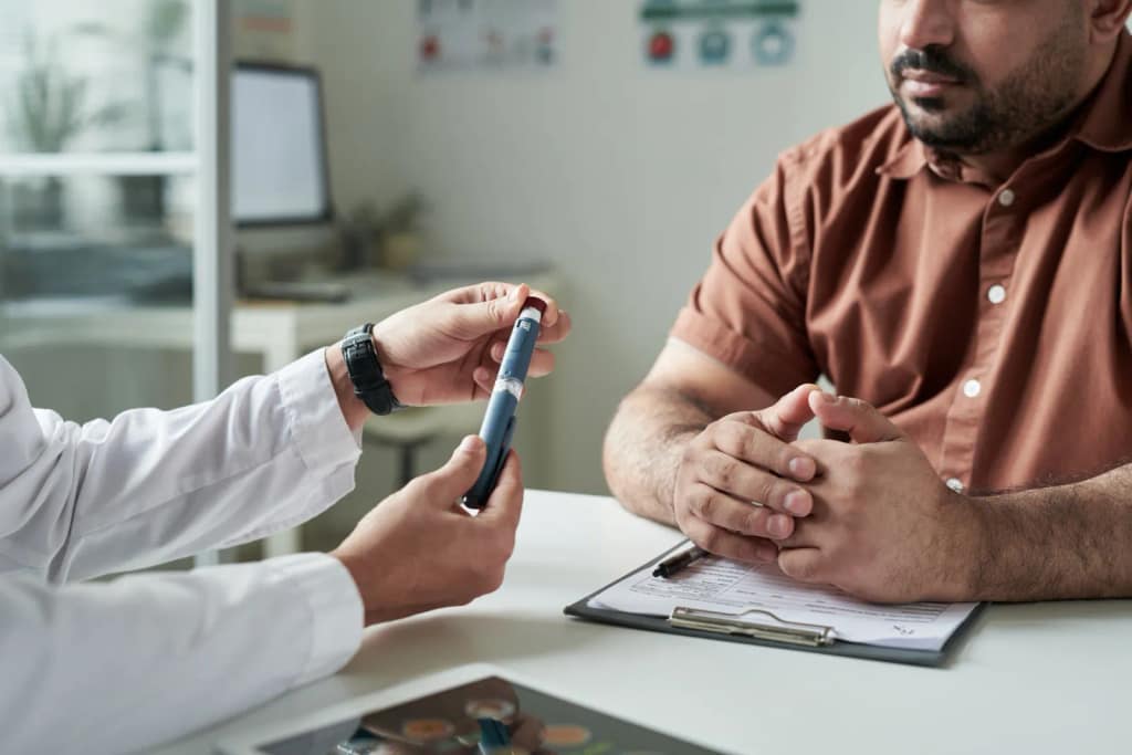 Image of a doctor and a patient in a consultation with no faces showing. Doctor holding a spring-loaded lancing device / Photo d'un médecin et d'un patient en consultation, dont les visages ne sont pas visibles. Le médecin tient un autopiqueur à ressort.