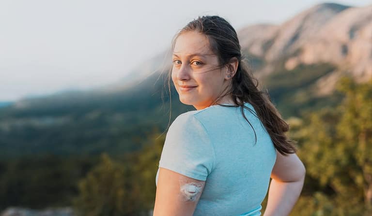 A young woman wearing a glucose sensor on her arm, mountains behind her