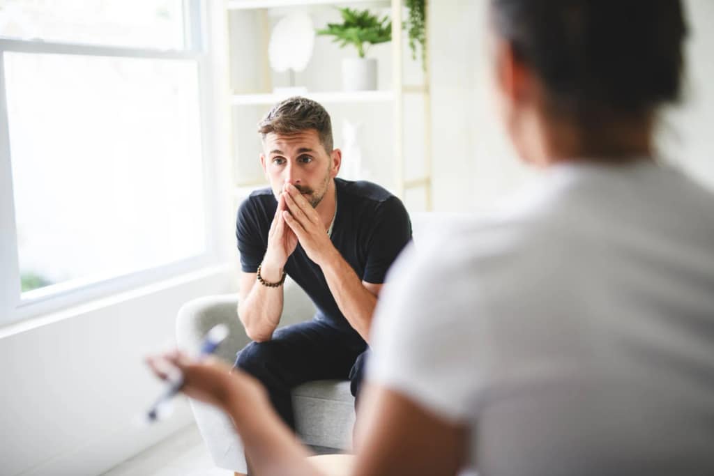 A young man sits on a chair with his hands clasped in front of his mouth; he looks pensive as he listens to an individual in the foreground.